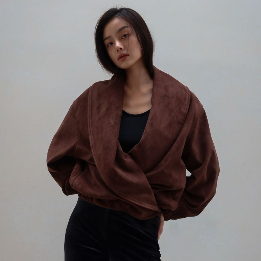 A model wearing a brown draped wrap jacket with a velvet-like texture, styled over a black top and black pants, standing against a plain light gray wall.