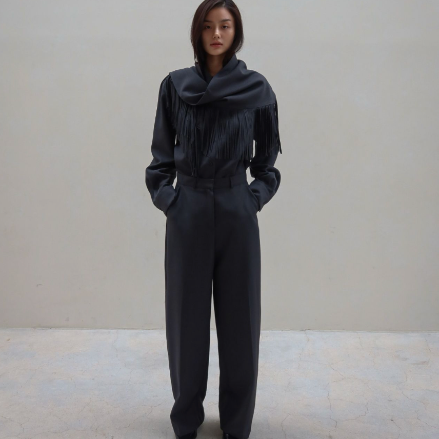 A model stands in a minimalist studio setting wearing a dark charcoal gray two-piece outfit consisting of a long-sleeve top with a dramatic draped collar and matching wide-leg trousers.