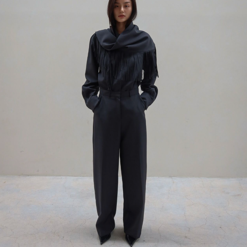 A model stands in a minimalist studio setting wearing a dark charcoal gray two-piece outfit consisting of a long-sleeve top with a dramatic draped collar and matching wide-leg trousers.