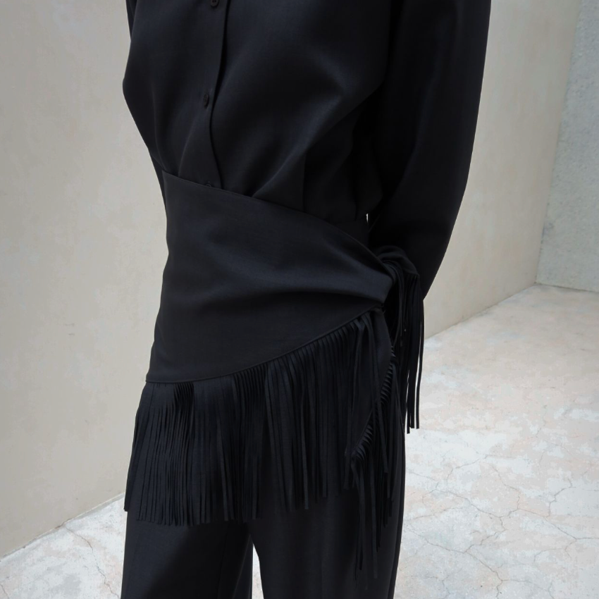 A person wearing a black satin-like top and wide-leg black pants with a long fringe detail at the waist, standing on a light stone floor against a beige wall.