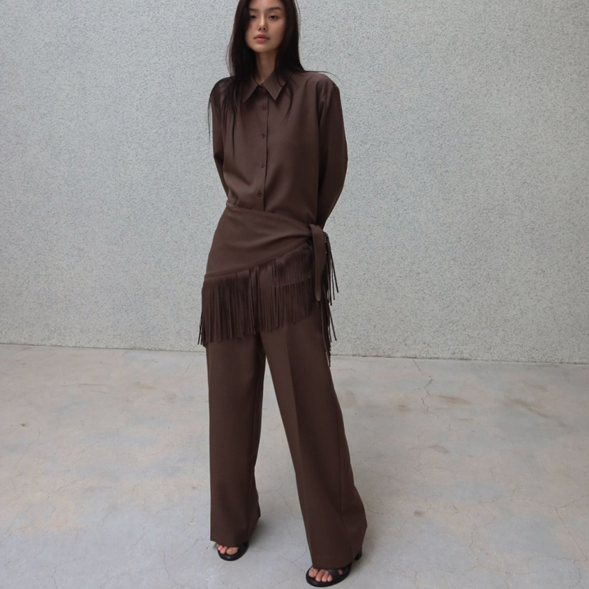 A model stands against a textured light gray wall wearing a matching brown two-piece outfit consisting of a long-sleeve collared shirt and wide-leg pants with fringe detailing.