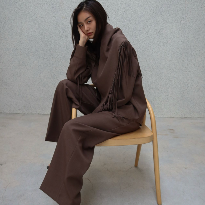 A model sits on a light wood chair wearing a matching dark brown lounge set consisting of a long-sleeve top with fringe details and wide-leg trousers, paired with black sandals, against a textured gray wall.