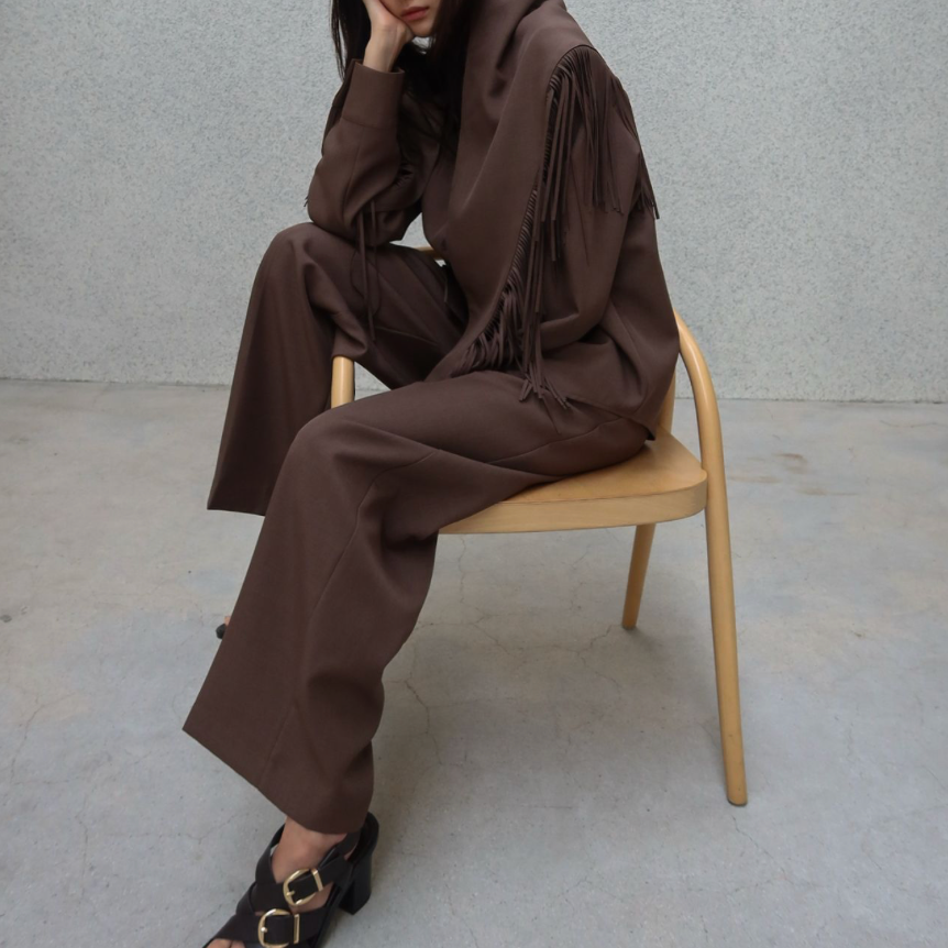 A model sits on a light wood chair wearing a matching dark brown lounge set consisting of a long-sleeve top with fringe details and wide-leg trousers, paired with black sandals, against a textured gray wall.