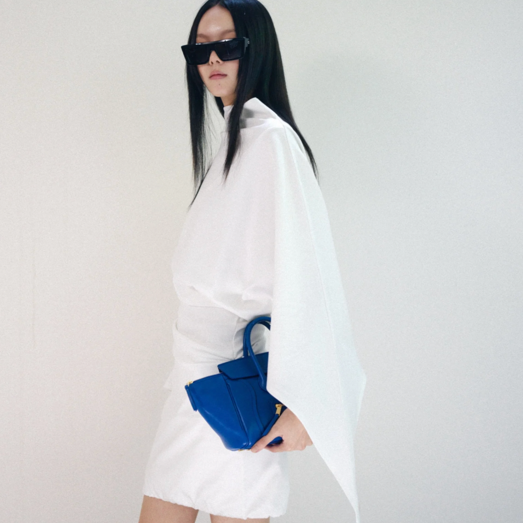 A model with long black hair and black sunglasses is shown from the side, wearing a white draped cape-style top and white shorts, holding a structured royal blue handbag against a plain off-white background.