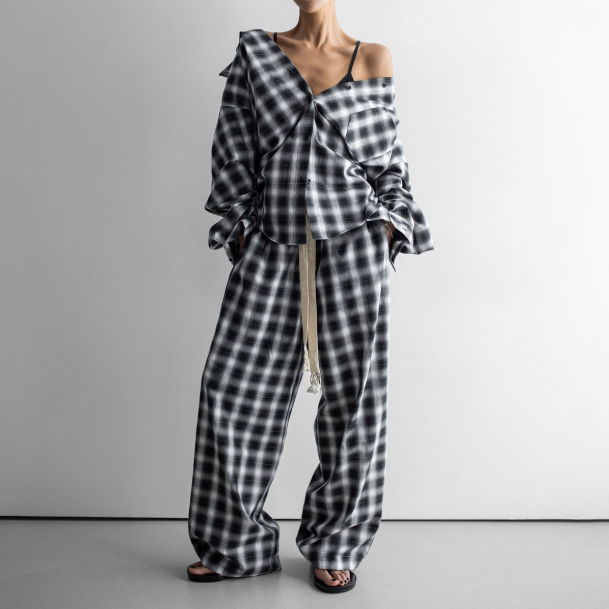 A model wearing a black and white checkered two-piece set consisting of an off-the-shoulder wrap top with ruffled sleeves and matching wide-leg pants with a drawstring waist, set against a minimalist studio background.