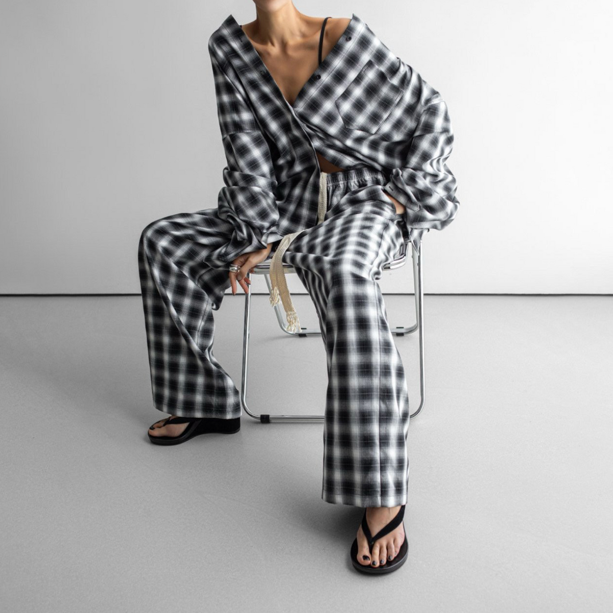 A model sits on a minimalist metal chair wearing a matching black and white gingham plaid lounge set, consisting of a long-sleeve wrap-style top and wide-leg trousers, paired with black flip-flop sandals.
