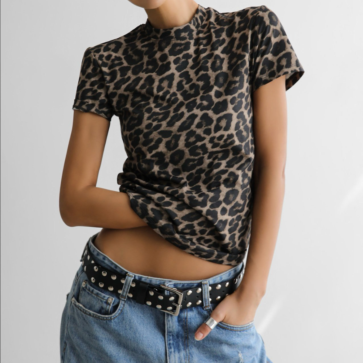 A person wearing a short-sleeve leopard-print top tucked into light-wash blue jeans with a black studded belt, photographed against a plain light background.