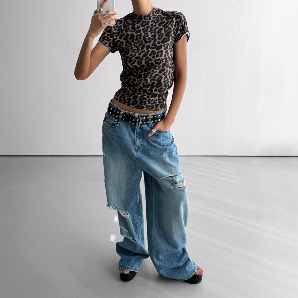 A model wearing a short-sleeve leopard-print crop top and distressed wide-leg blue jeans with a studded black belt, standing in a minimalist studio setting.