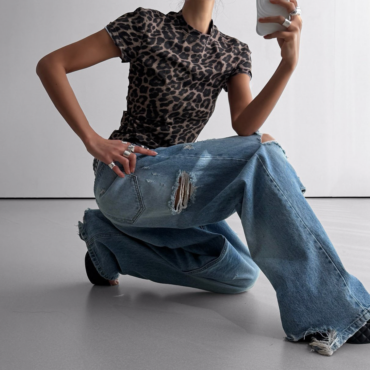 A model wearing a short-sleeve leopard-print t-shirt and wide-leg distressed blue jeans, kneeling on a light gray floor against a white wall.