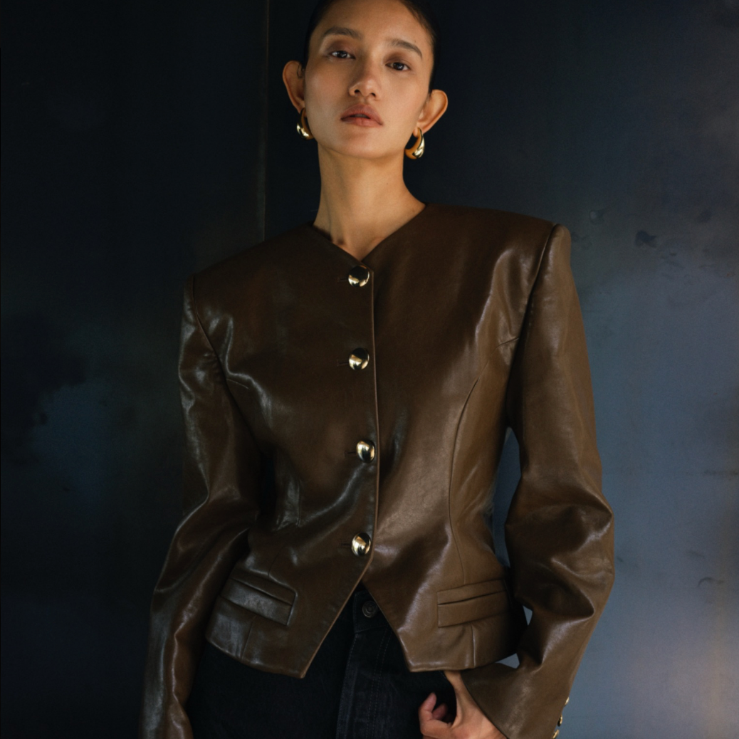 A woman wearing a cropped brown faux-leather jacket with gold-tone buttons, styled with black pants against a dark textured studio background.