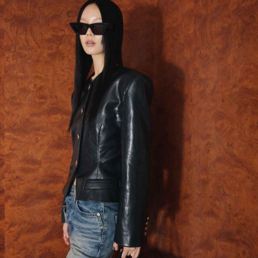 A model with long black hair wears a black leather jacket, black sunglasses, and blue jeans, standing against a textured orange-brown wall and holding a black bag.
