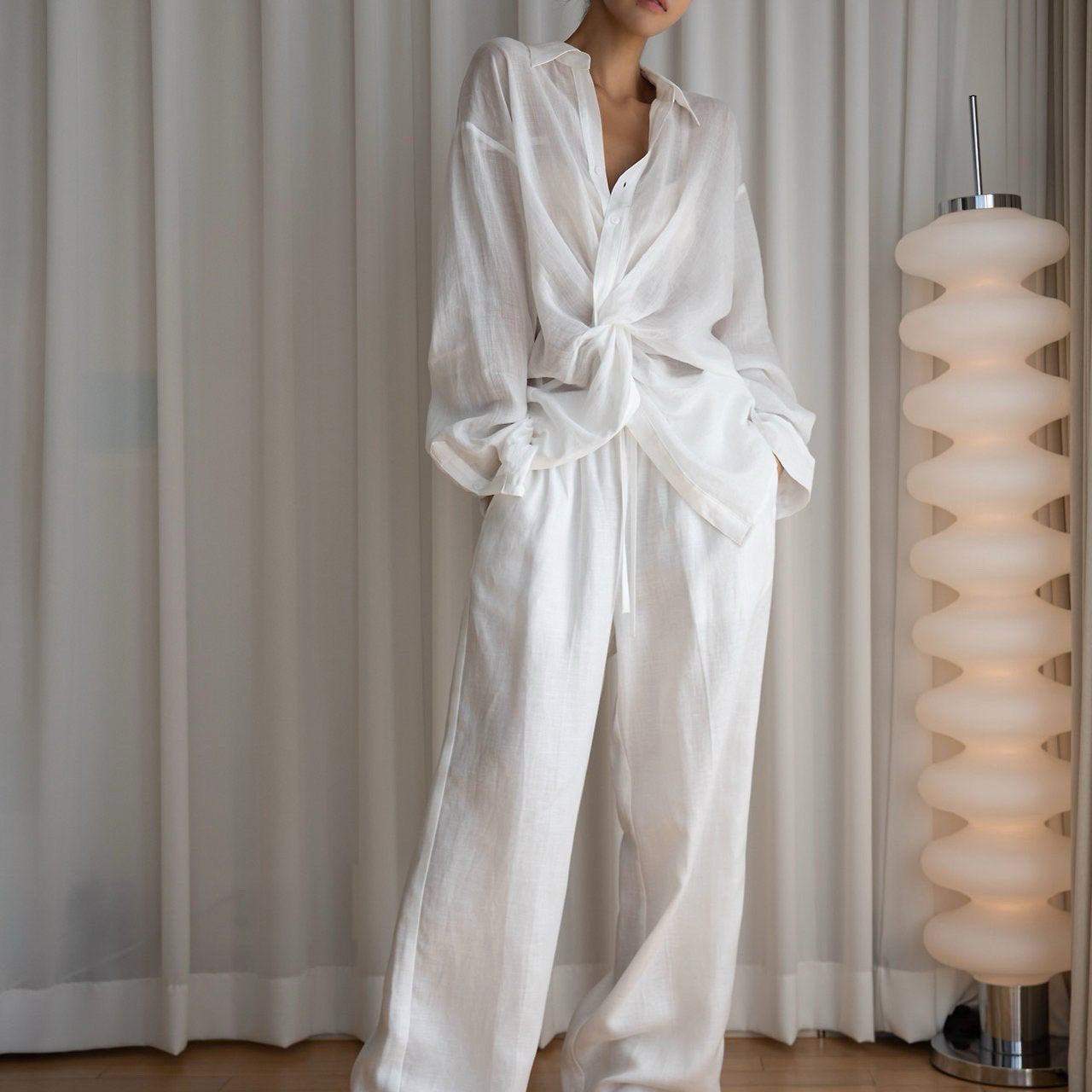 Person wearing a white satin robe standing in a room with light-colored curtains and a lamp.