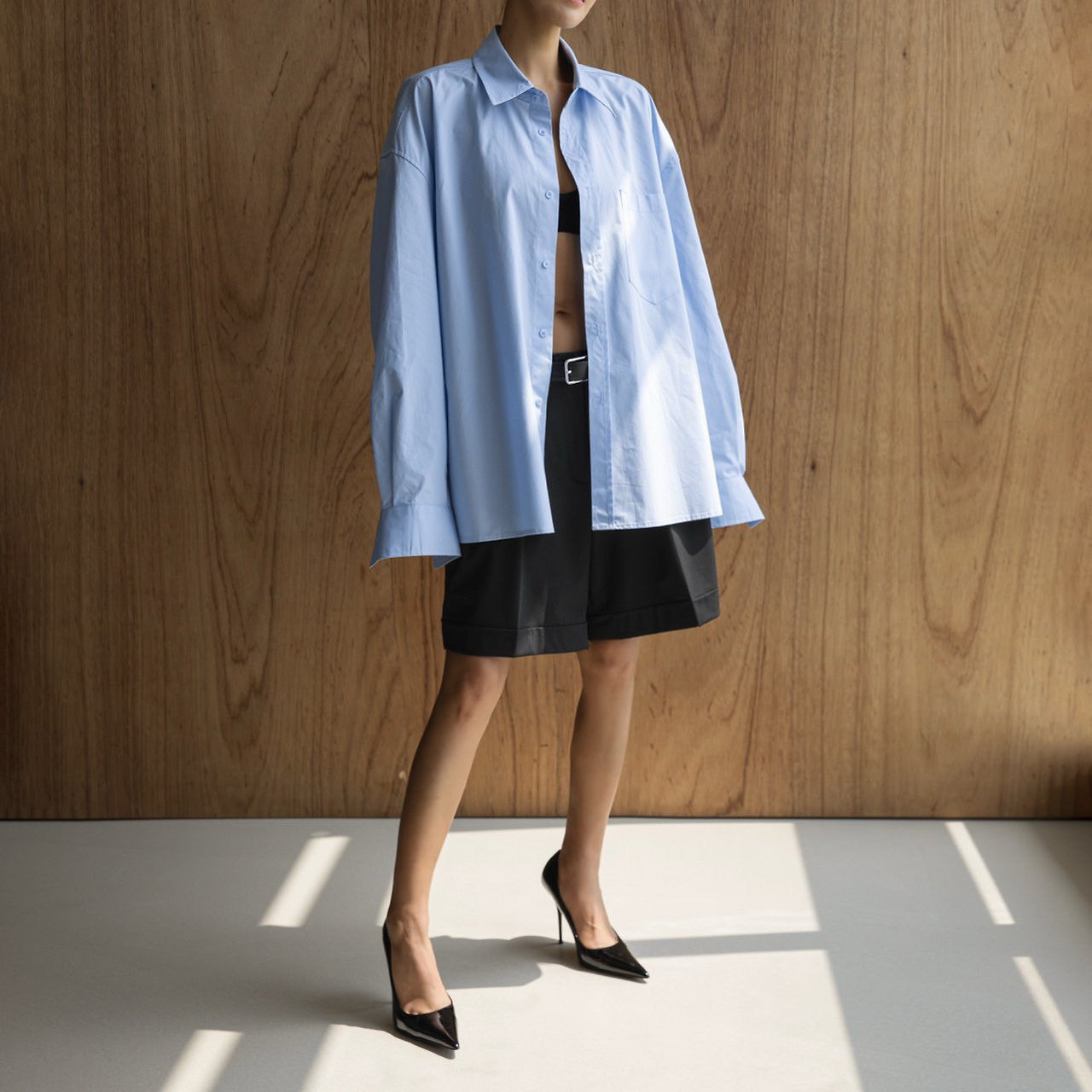 Person wearing a light blue shirt over a black top and skirt, standing against a wooden wall.