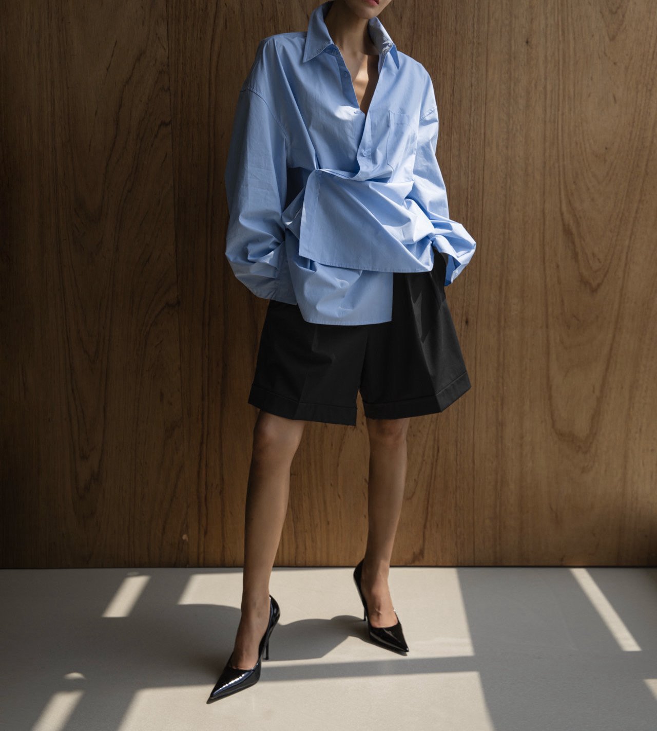 Person wearing a light blue oversized shirt and black shorts against a wooden wall.