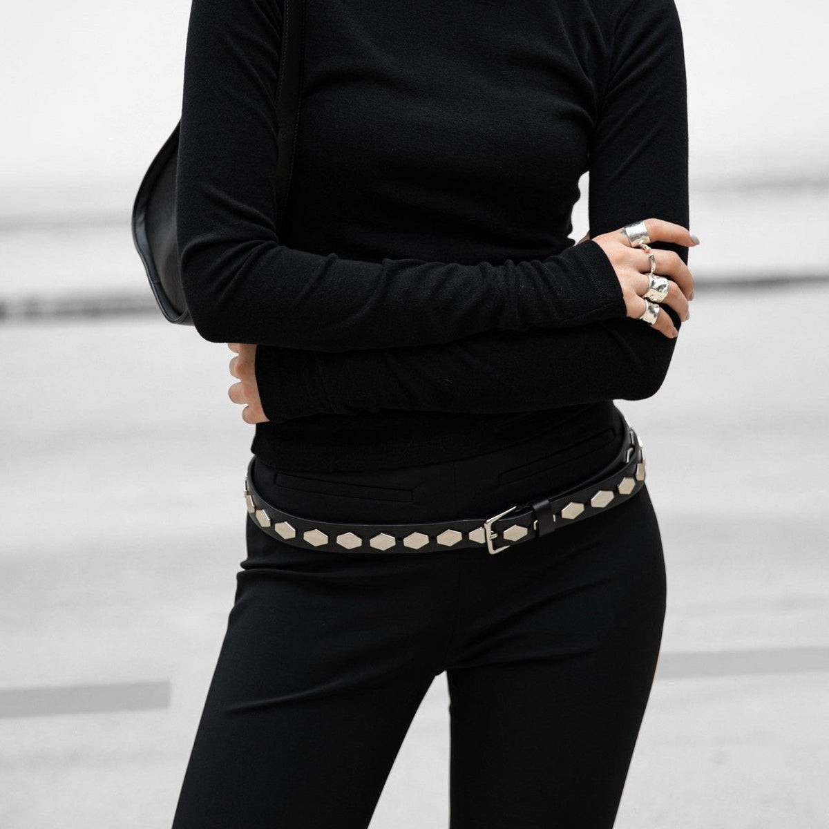 Person wearing a black turtleneck and black pants with a decorative belt against a light background