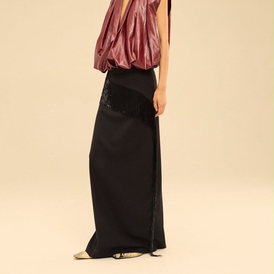 A model wearing a dark burgundy draped sleeveless top and a long black skirt with fringe detailing, standing against a solid beige studio background.