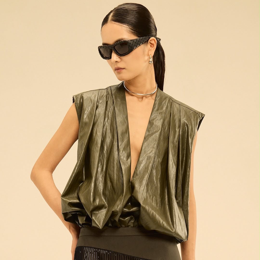 A model wearing a metallic olive green draped sleeveless top and a matching dark fringe skirt, styled with black sunglasses and a choker necklace against a solid beige background.