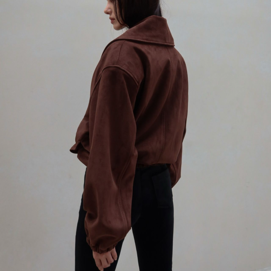 A woman wearing a cropped brown suede jacket and black pants, photographed from the side against a plain beige wall.