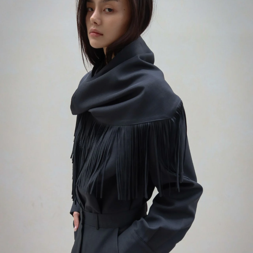 A model wearing a black long-sleeve top with a wide draped collar and cascading fringe, paired with matching black trousers, set against a plain light beige wall.