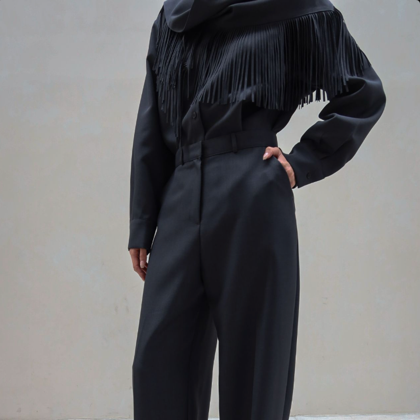 A model wearing a black long-sleeve top with a fringe yoke and matching black wide-leg trousers, paired with black pointed-toe heels, standing against a neutral beige wall.