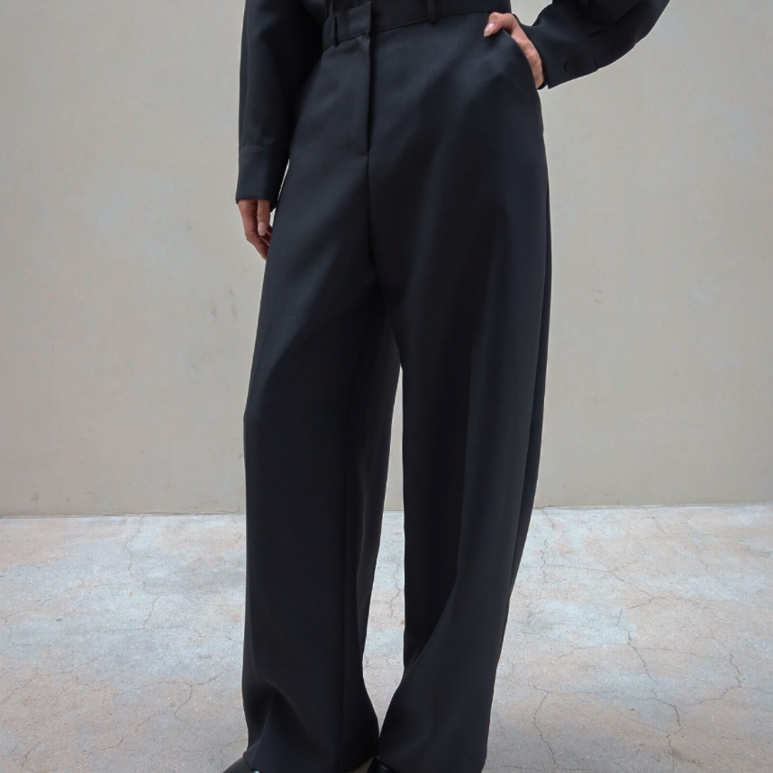A model wearing a black long-sleeve top with a fringe yoke and matching black wide-leg trousers, paired with black pointed-toe heels, standing against a neutral beige wall.