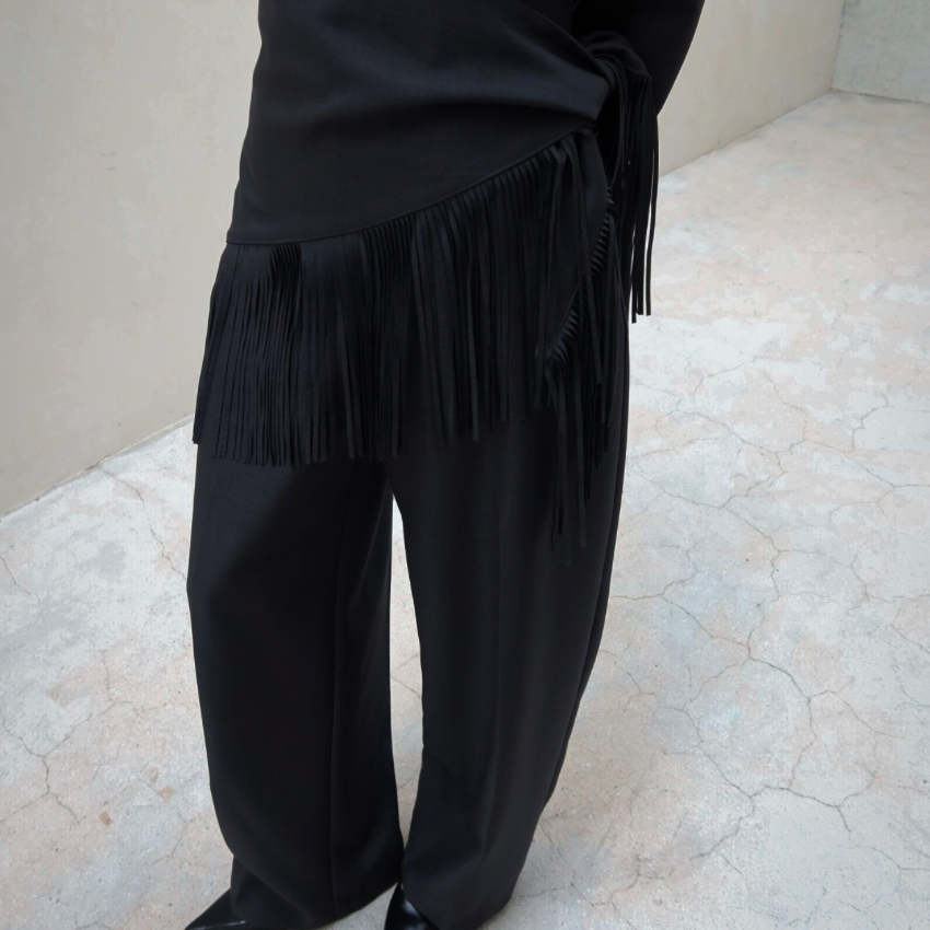 A person wearing a black satin-like top and wide-leg black pants with a long fringe detail at the waist, standing on a light stone floor against a beige wall.