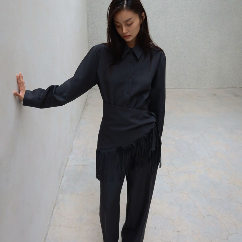 A woman wearing a black long-sleeve collared shirt with fringe detailing at the hem and matching black wide-leg trousers, standing in a minimalist gray room and touching the wall with her left hand.