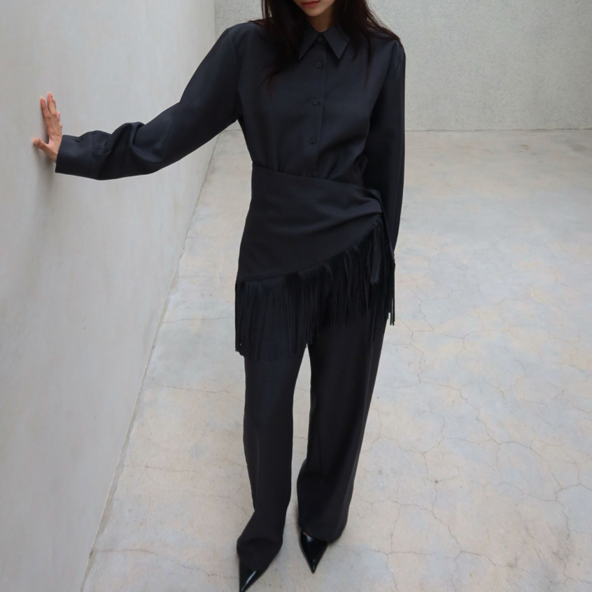 A woman wearing a black long-sleeve collared shirt with fringe detailing at the hem and matching black wide-leg trousers, standing in a minimalist gray room and touching the wall with her left hand.