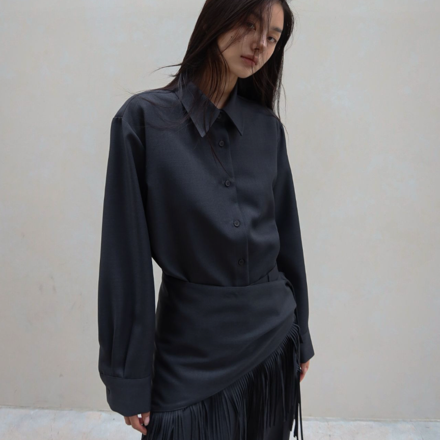 A woman wearing a dark charcoal gray long-sleeve button-up shirt and a matching dark fringe skirt, standing against a textured light beige wall.