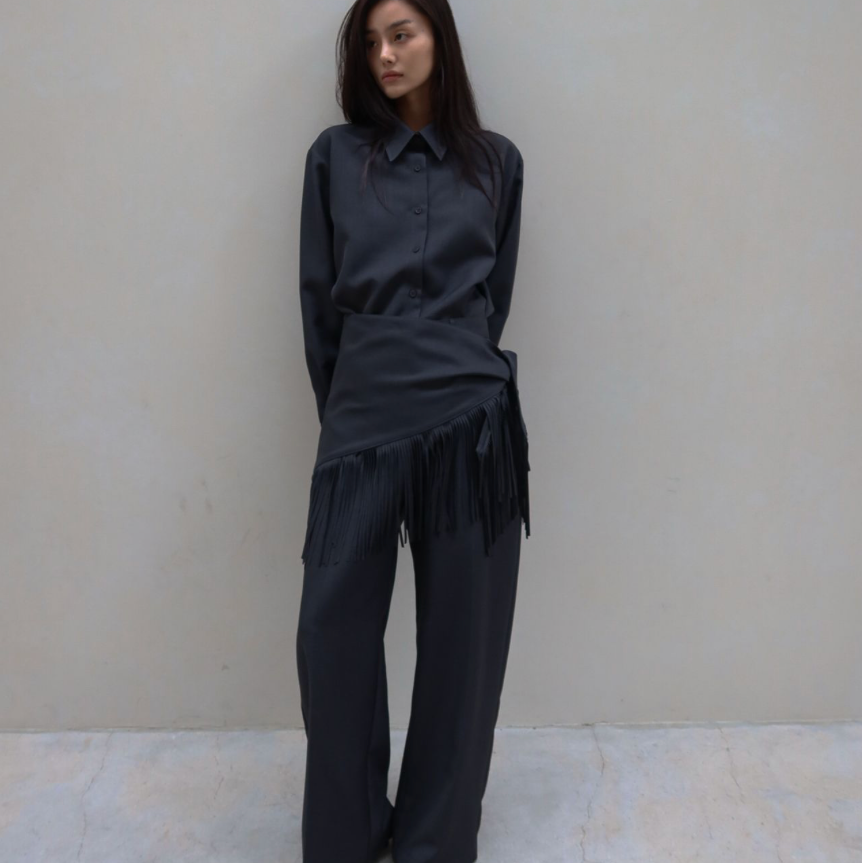 A model stands against a textured beige wall wearing a dark charcoal gray matching set consisting of a long-sleeve collared shirt with fringe detailing at the waist and matching wide-leg trousers.