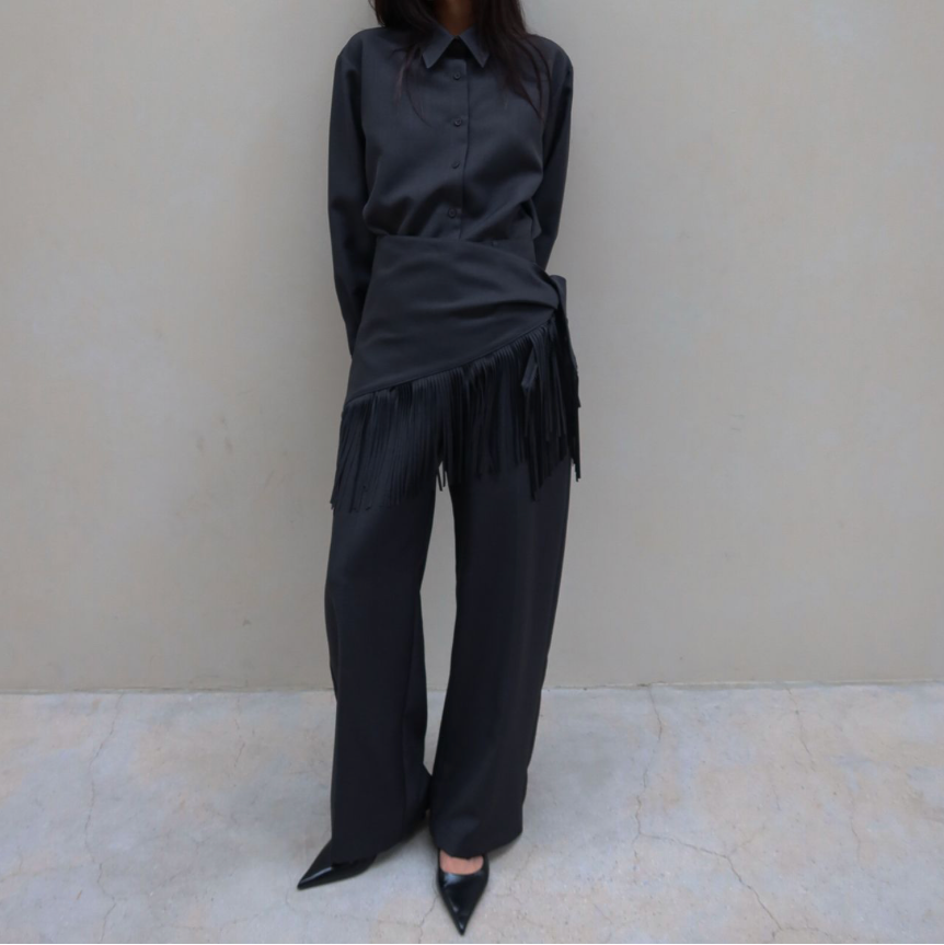 A model stands against a textured beige wall wearing a dark charcoal gray matching set consisting of a long-sleeve collared shirt with fringe detailing at the waist and matching wide-leg trousers.