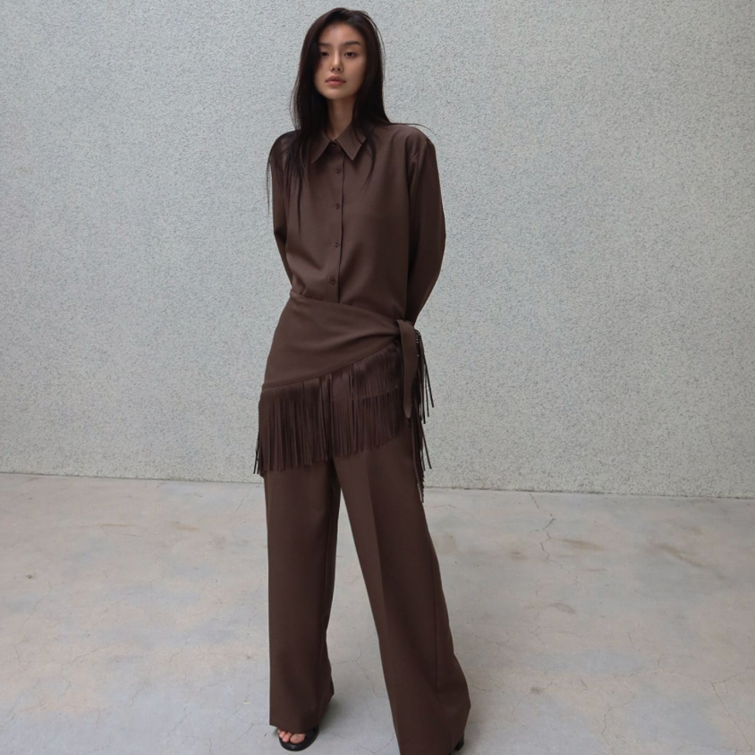 A model stands against a textured light gray wall wearing a matching brown two-piece outfit consisting of a long-sleeve collared shirt and wide-leg pants with fringe detailing.