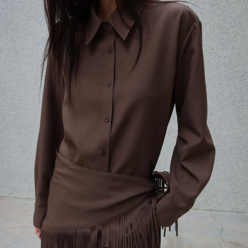 A person wearing a brown long-sleeve collared button-front top with a draped wrap-style hem and fringe detail, paired with matching brown pants, against a light gray textured wall.