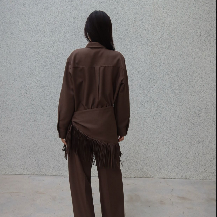 A person viewed from behind wearing a brown long-sleeve collared top with fringe detail and matching wide-leg pants, standing against a light gray textured wall.