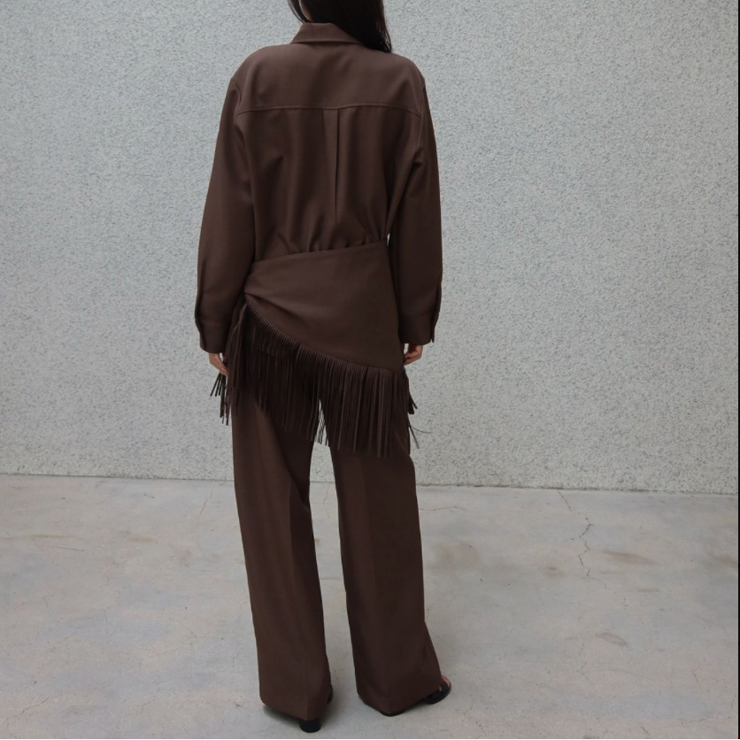 A person viewed from behind wearing a brown long-sleeve collared top with fringe detail and matching wide-leg pants, standing against a light gray textured wall.