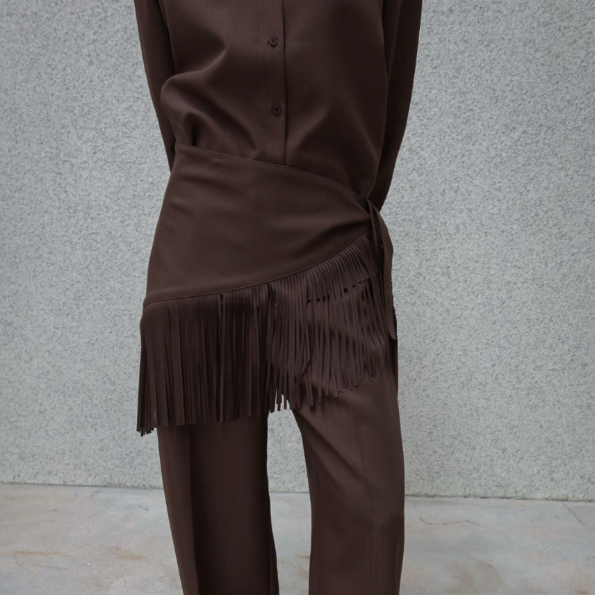 A person wearing a matching brown satin-like button-up shirt and wide-leg pants set, with the pants featuring a draped fringed waist detail. The model is standing against a textured gray wall and concrete floor.