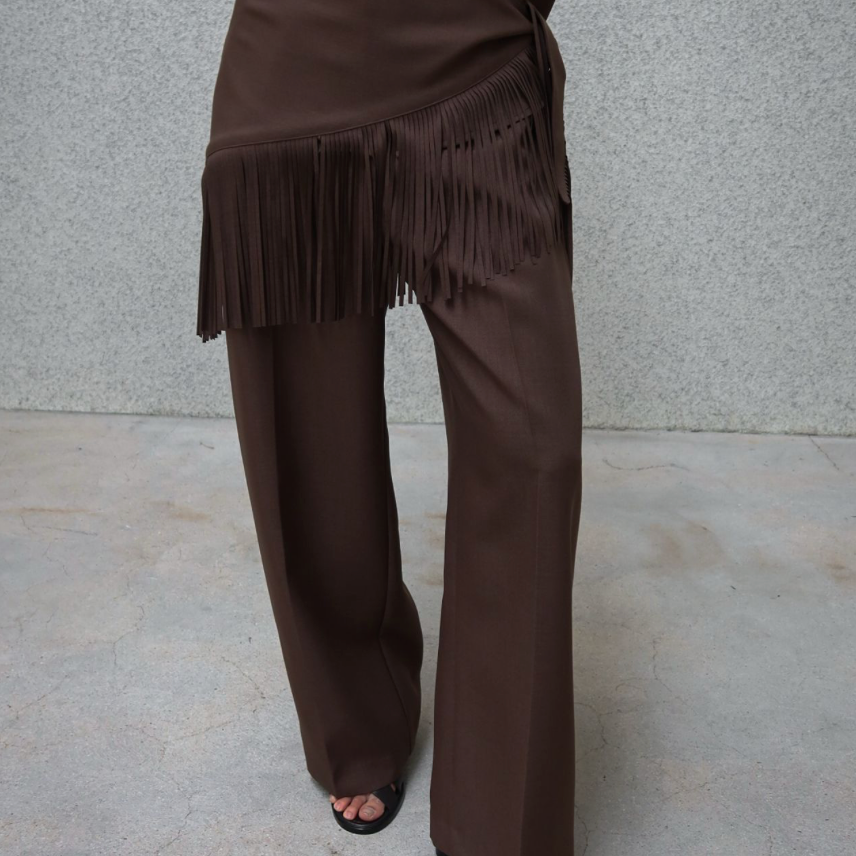 A person wearing a matching brown satin-like button-up shirt and wide-leg pants set, with the pants featuring a draped fringed waist detail. The model is standing against a textured gray wall and concrete floor.