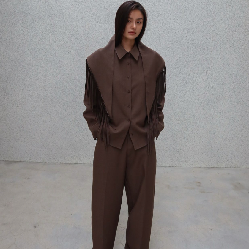 A model stands against a textured light gray wall wearing a matching brown wide-leg suit with a fringed shawl-style collar and a button-front jacket.