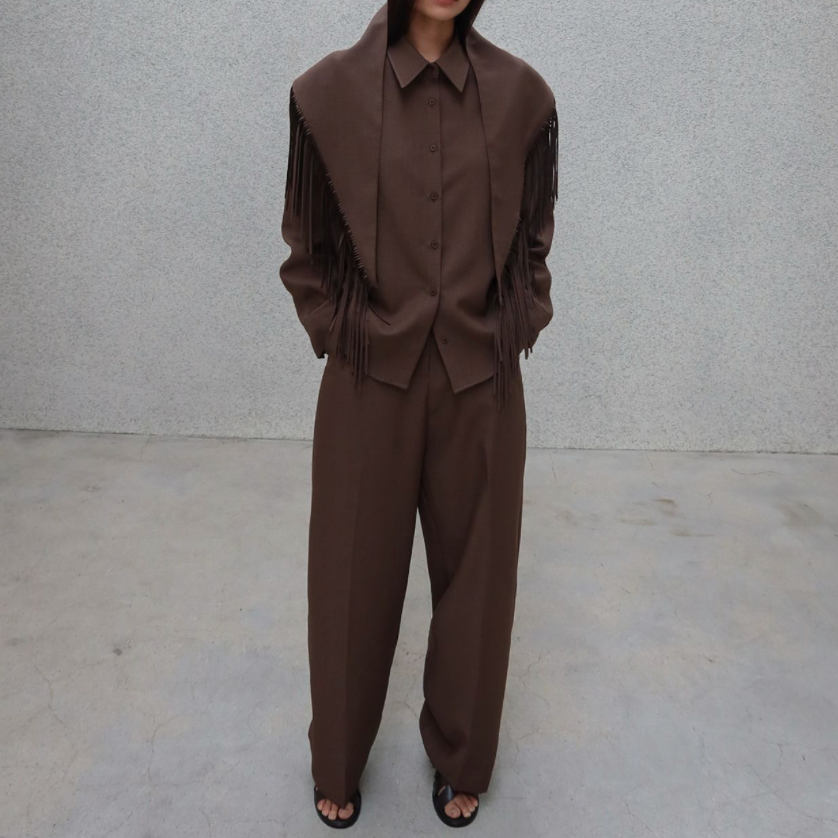 A model stands against a textured light gray wall wearing a matching brown wide-leg suit with a fringed shawl-style collar and a button-front jacket.