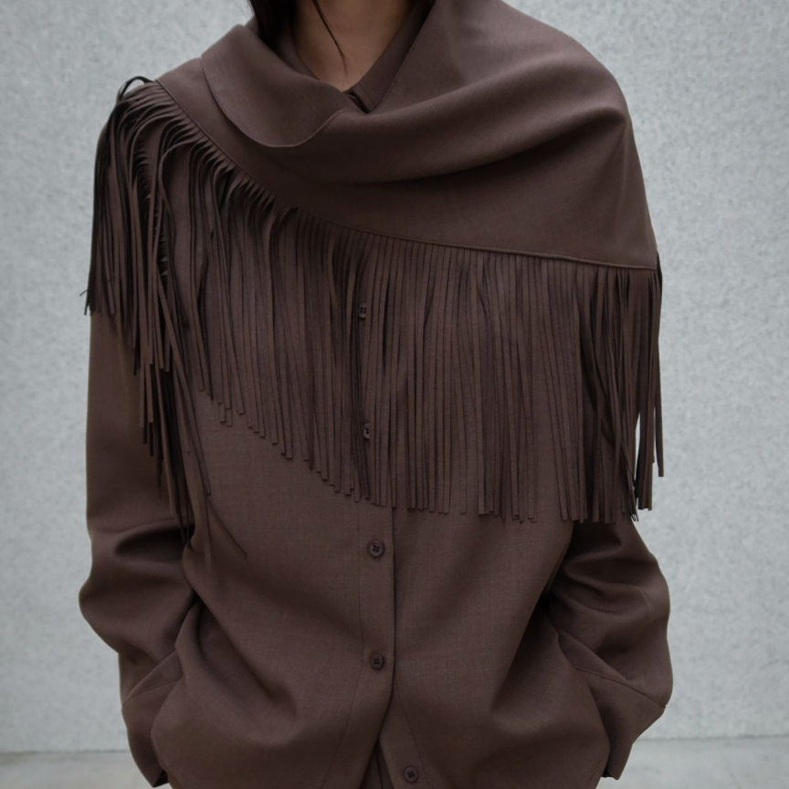 A person wearing a brown fringed shawl draped over a matching brown button-front top and trousers, photographed against a light gray textured wall.