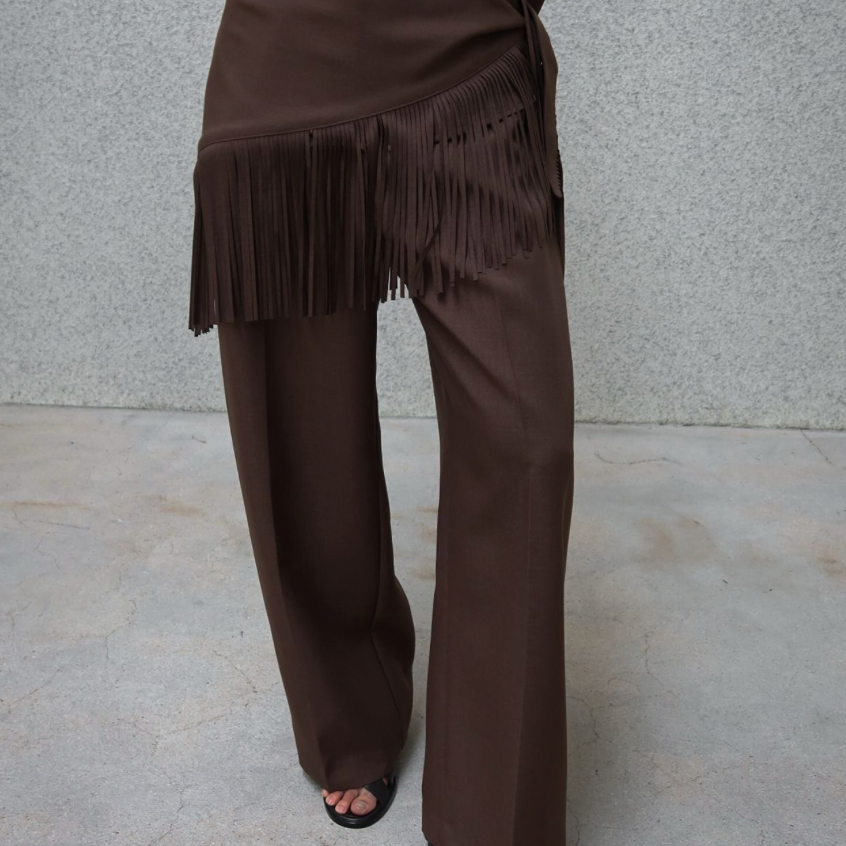 A person wearing a dark brown matching set consisting of wide-leg trousers and a long-sleeve top with a fringed wrap detail at the waist, paired with black slide sandals, standing against a textured gray wall.