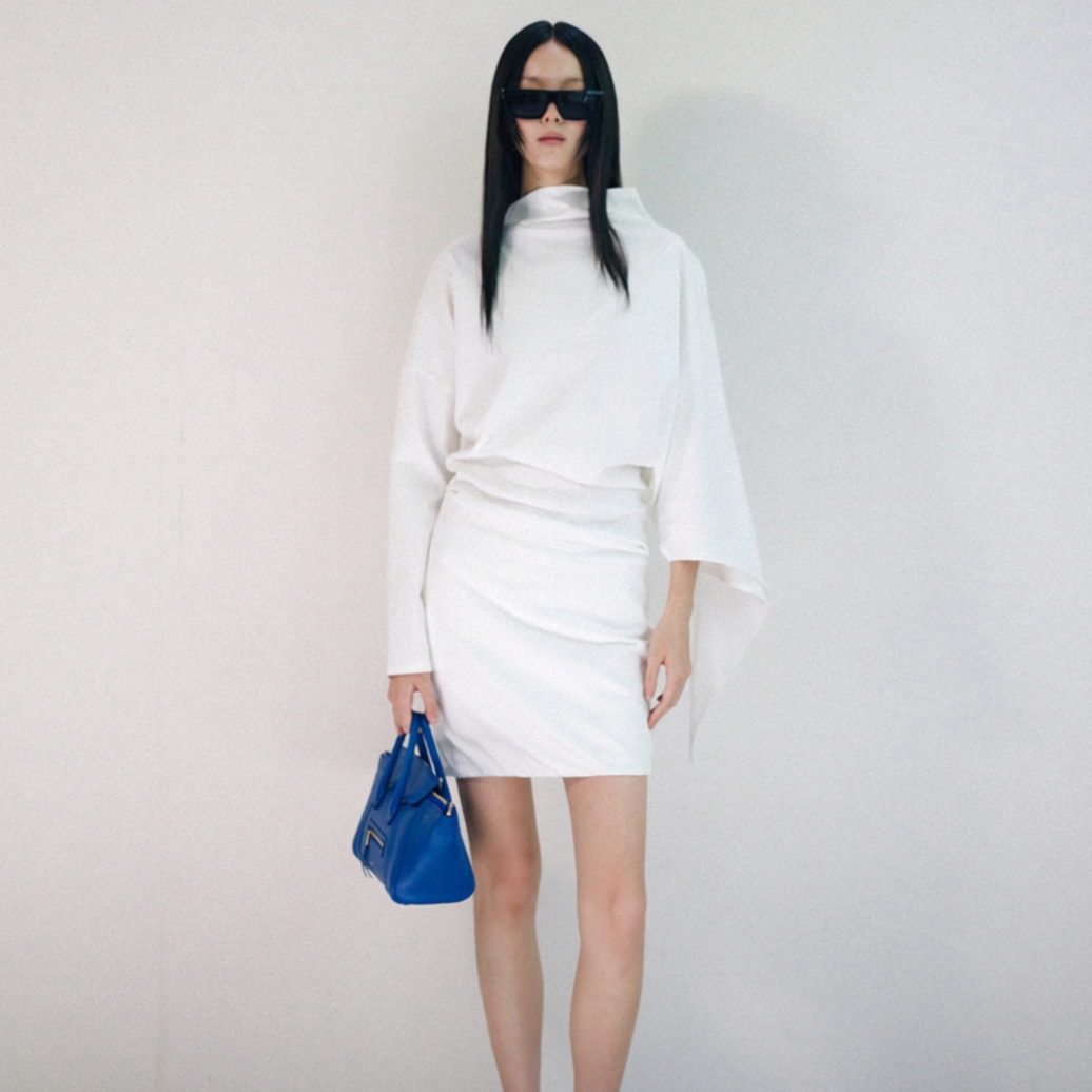 A model with long black hair and sunglasses wearing a white long-sleeve mock-neck top with draped sleeves and a matching white ruched mini skirt, holding a blue handbag and wearing black loafers, standing against a plain off-white wall.