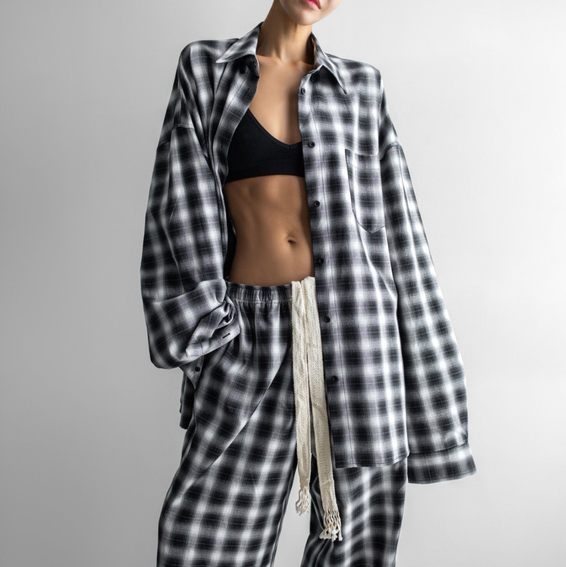 A model wearing a matching black and white plaid lounge set, featuring an oversized long-sleeve button-up shirt and matching pants with a cream drawstring, styled over a black sports bra against a light gray background.