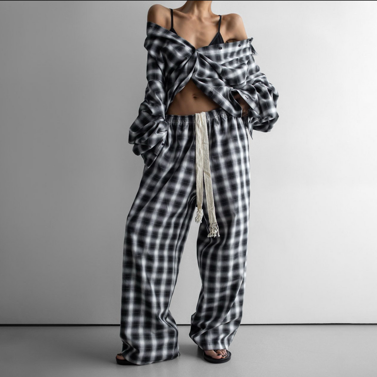 A model wearing a matching black and white plaid lounge set, featuring an off-the-shoulder long-sleeve top and wide-leg pants with a cream drawstring, set against a plain gray studio background.