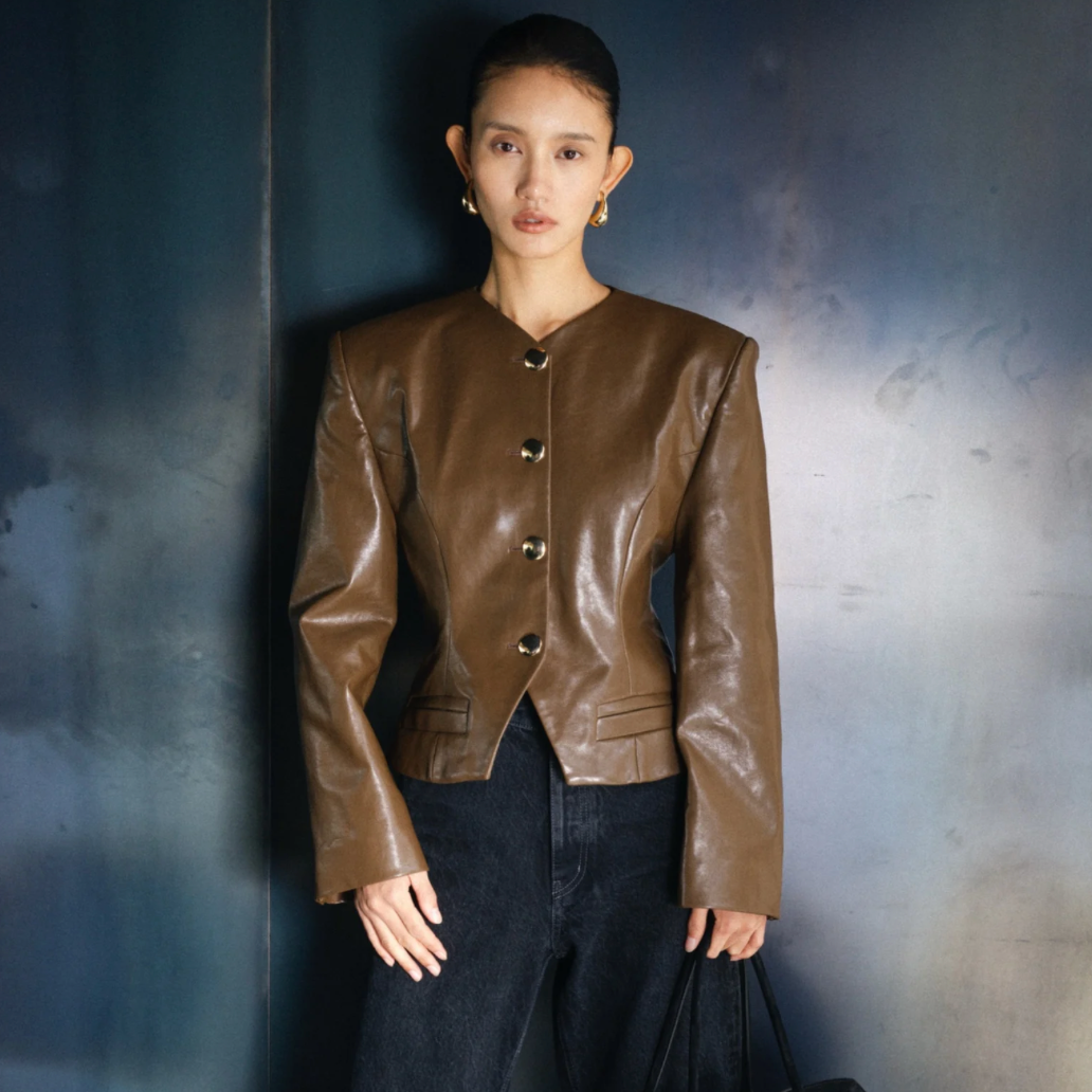 A model stands against a textured dark blue-gray wall wearing a cropped brown faux-leather jacket with a button front, paired with dark jeans and holding a black tote bag.