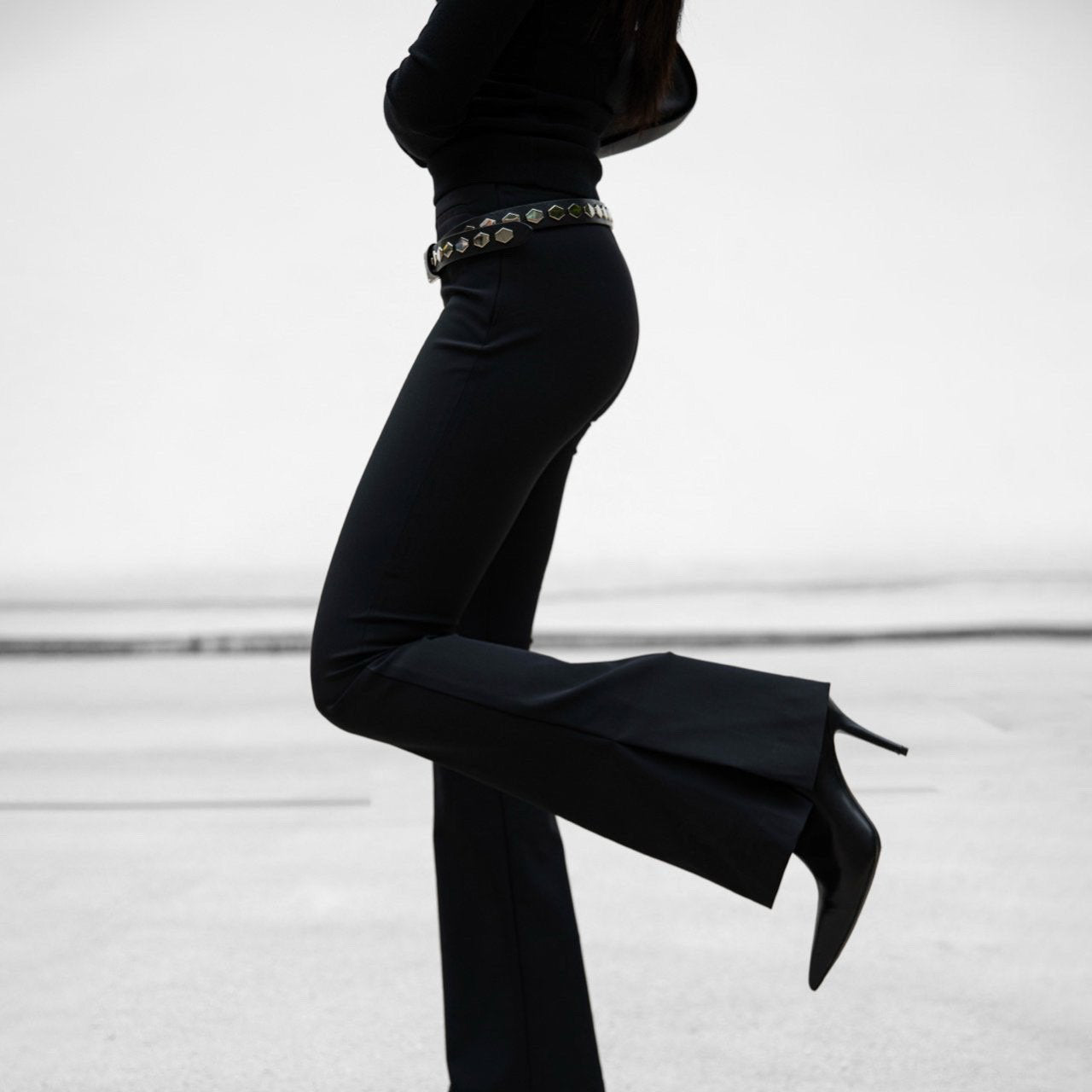 Person wearing a black outfit with high-waisted pants and black shoes on a light background